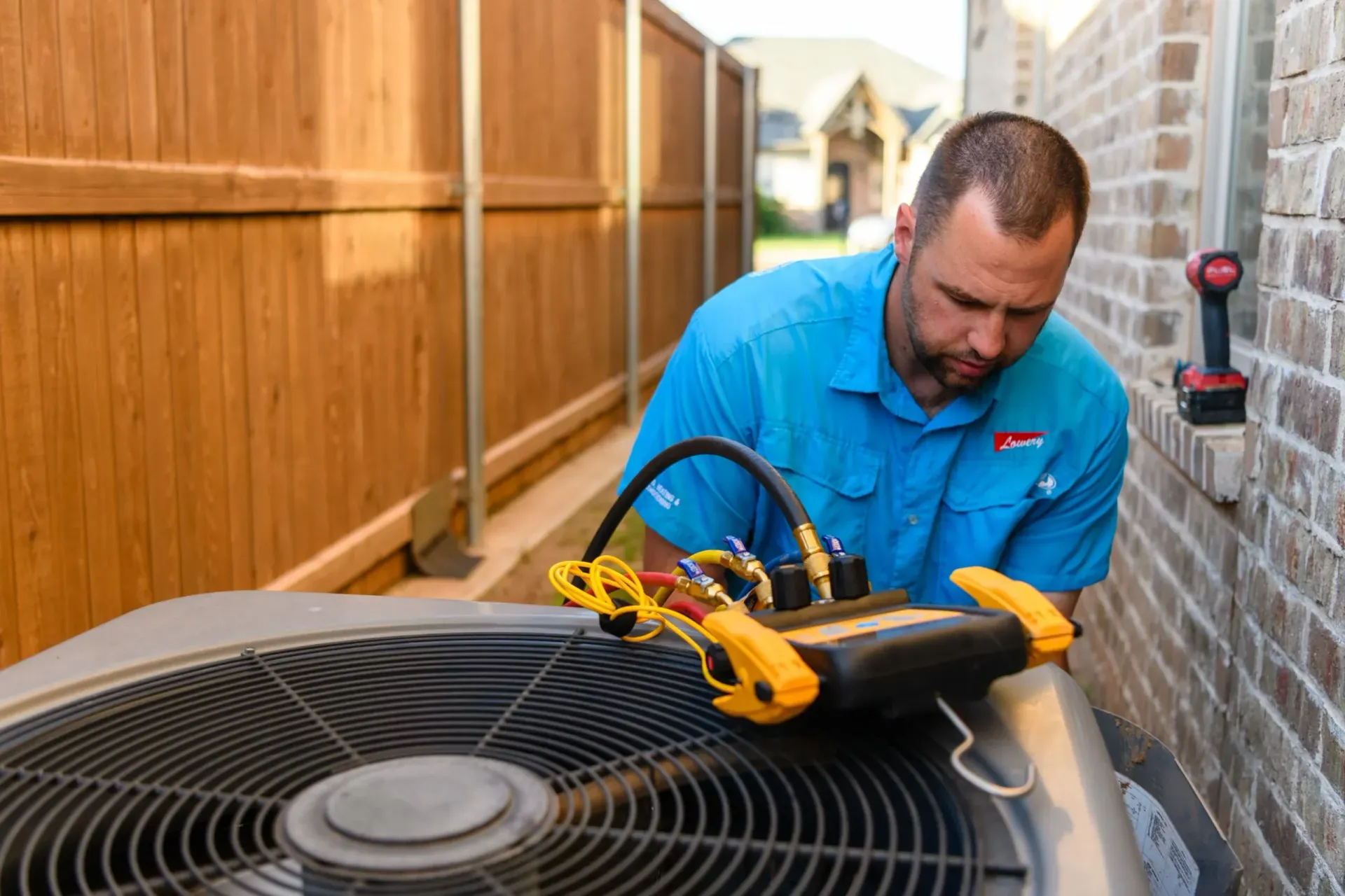 HVAC technician repairs an air conditioning unit outside a brick wall and wooden fence.