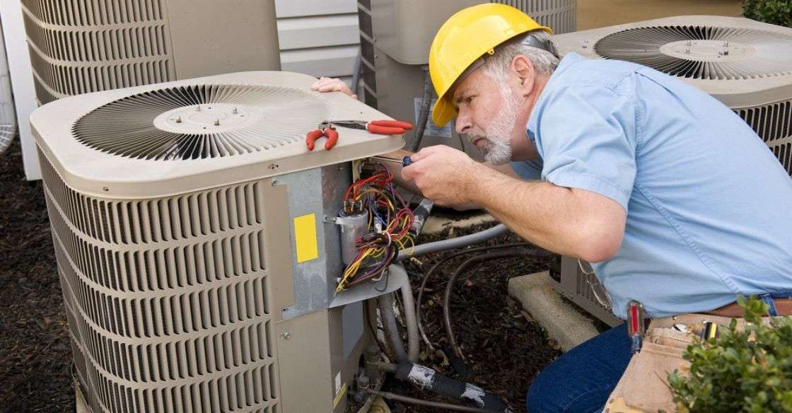 Technician in hard hat repairing an air conditioner unit outdoors, using tools.