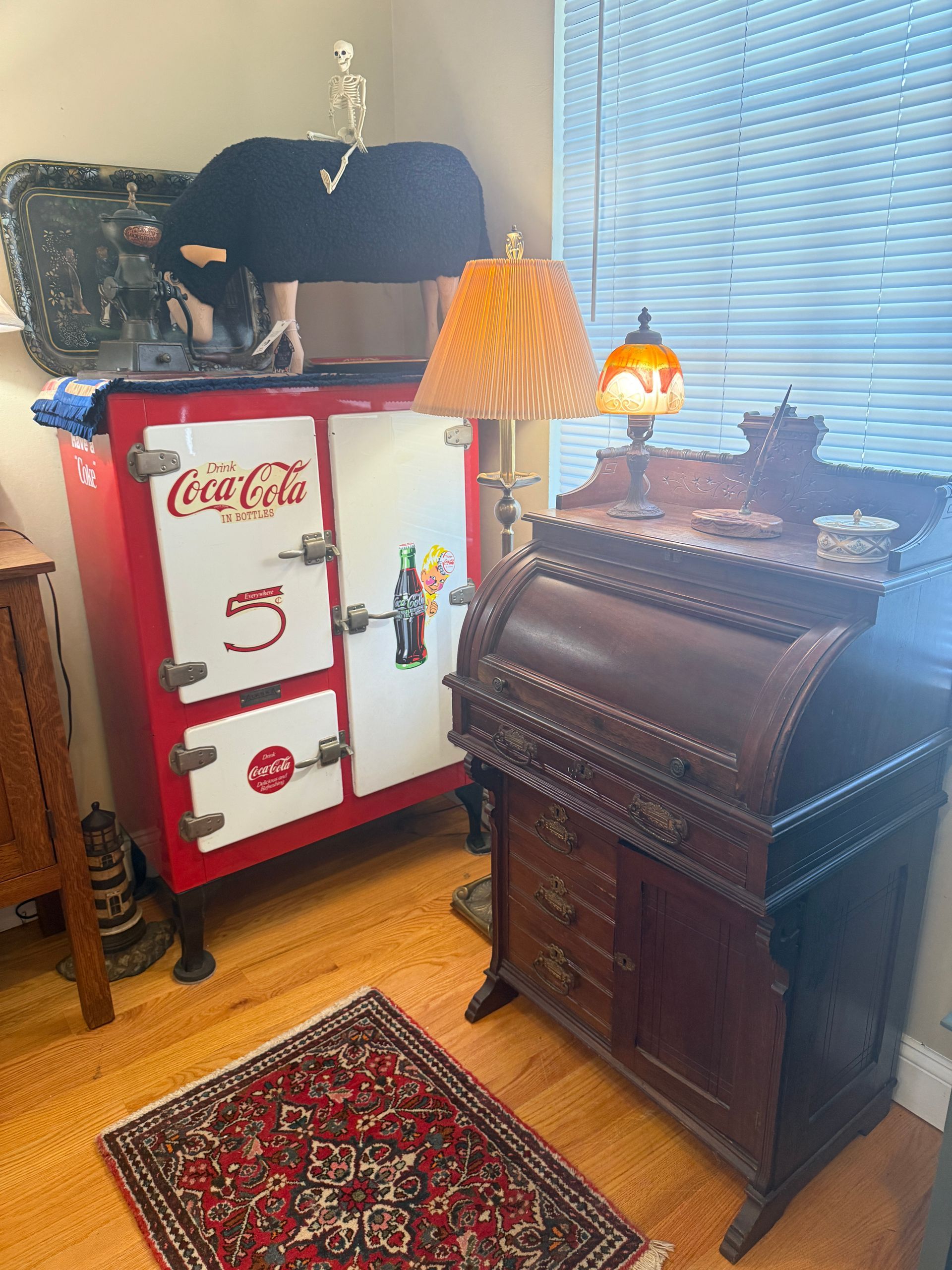 Red and white Coca-Cola cooler, antique desk, lamps, and rug in a room with a window.