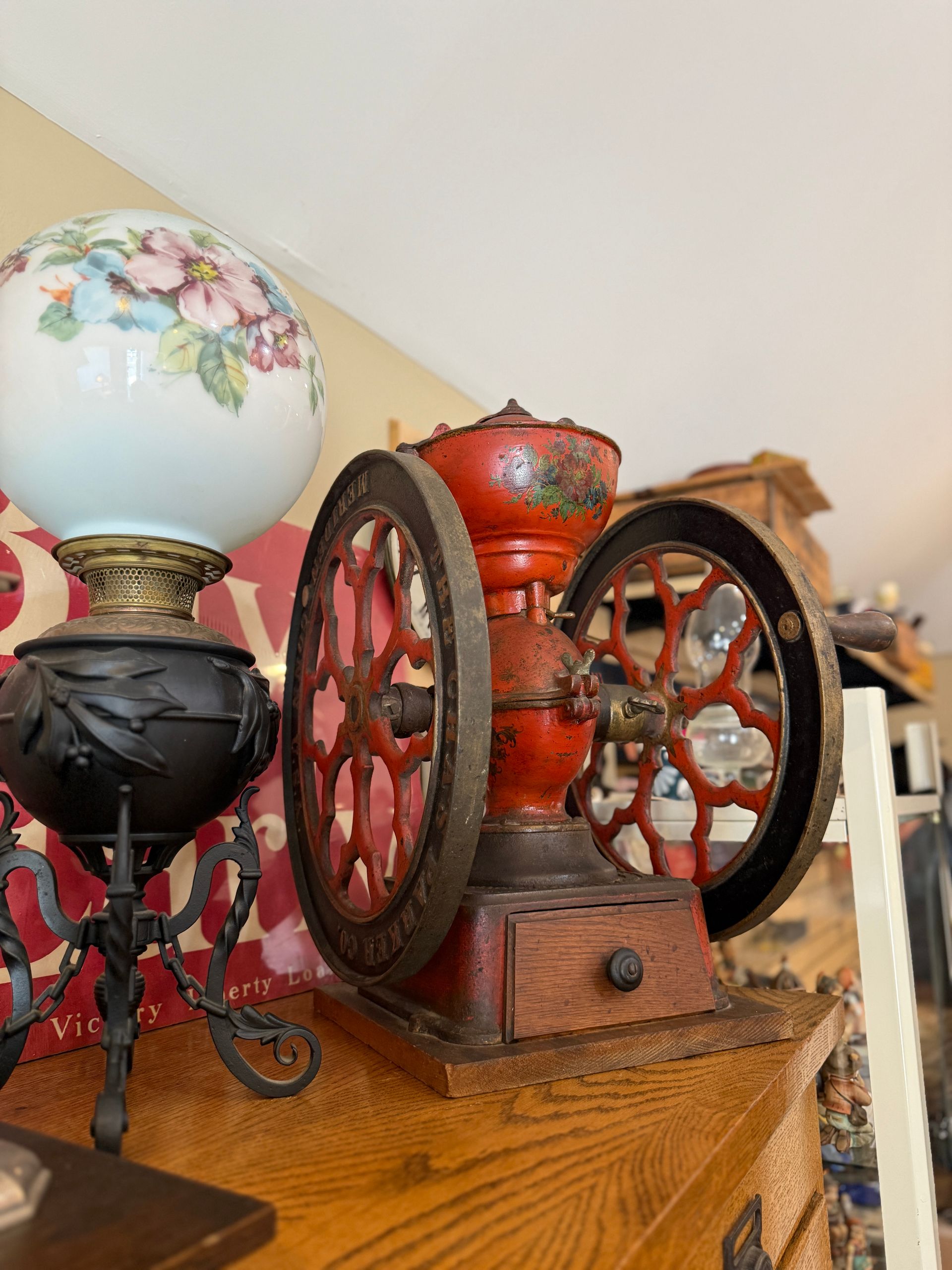 Antique red coffee grinder with large spoked wheels, next to a decorative lamp with a floral globe.