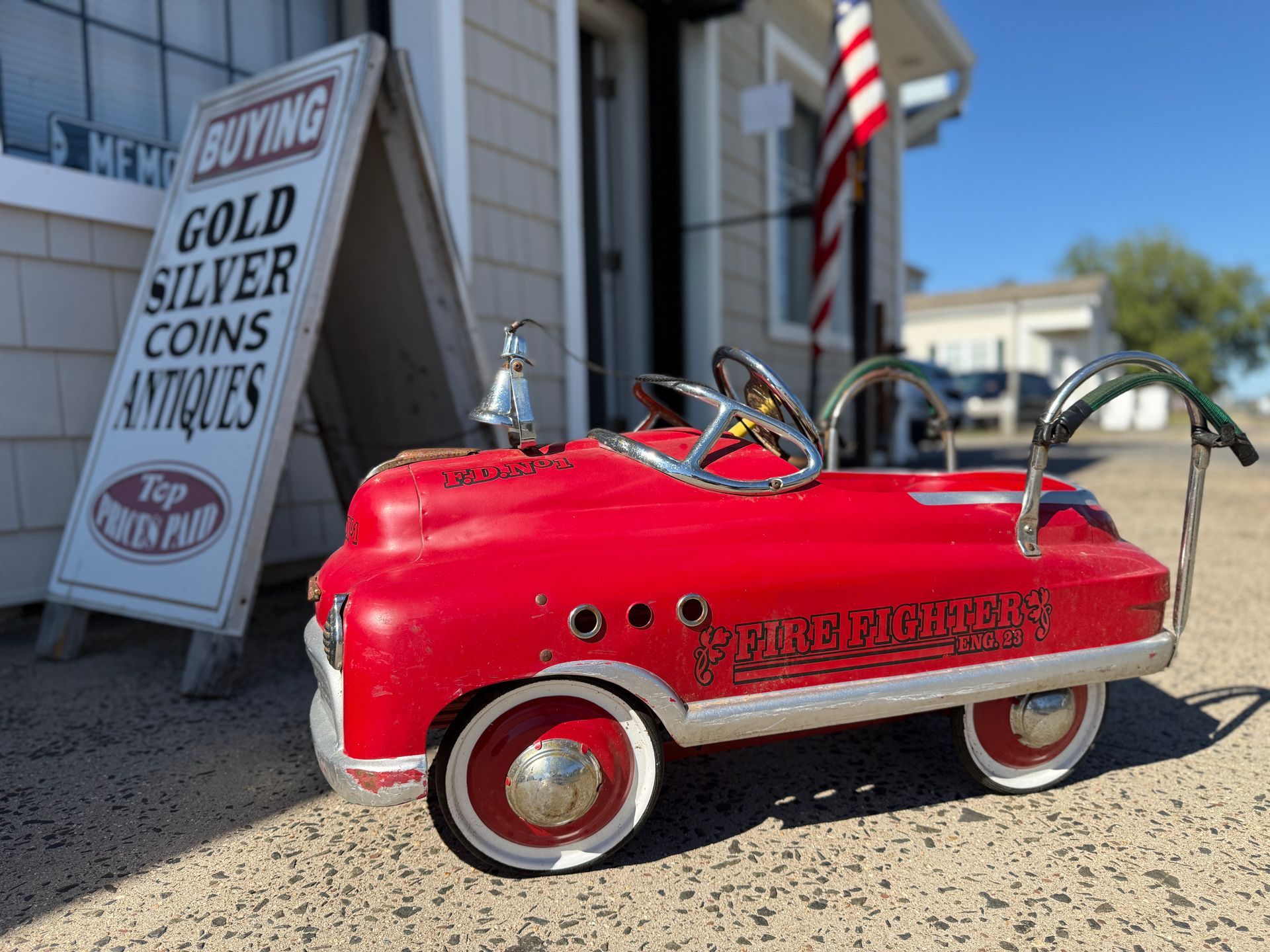 Red pedal car parked outside of Ricks Antiques & Coins shop advertising to buy gold, silver coins, and antiques.