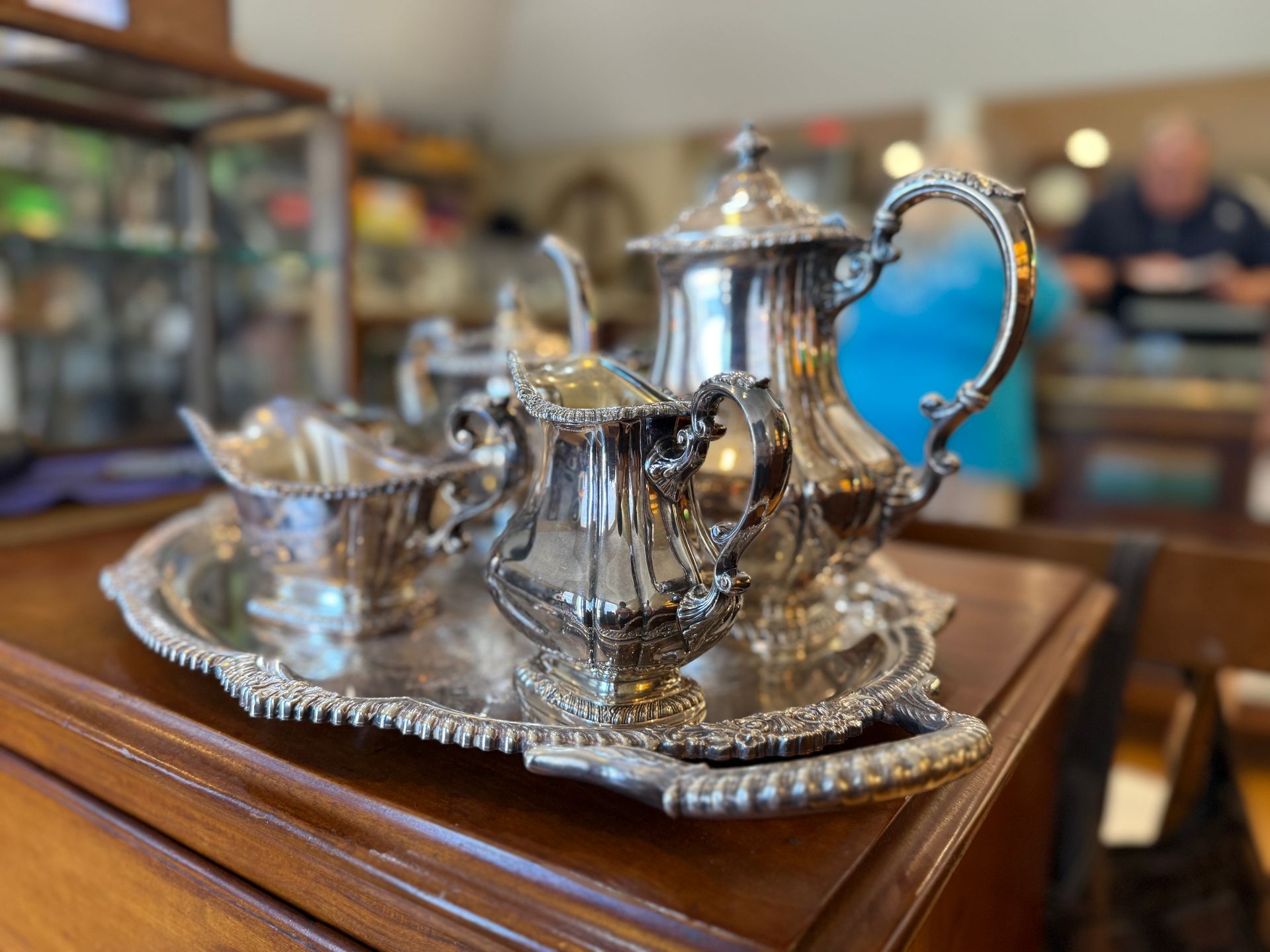 Silver tea set on a decorative tray, set on a wooden surface, with a blurry interior background.