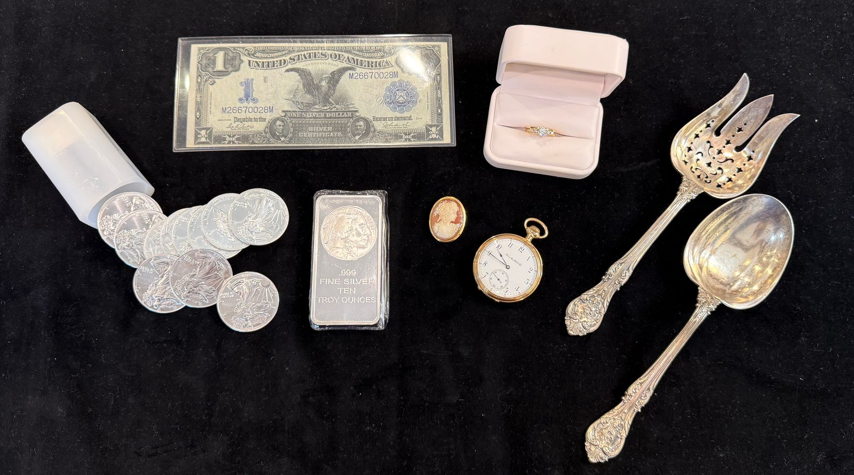 Silver coins, bar, and assorted jewelry with a dollar bill on a black surface.