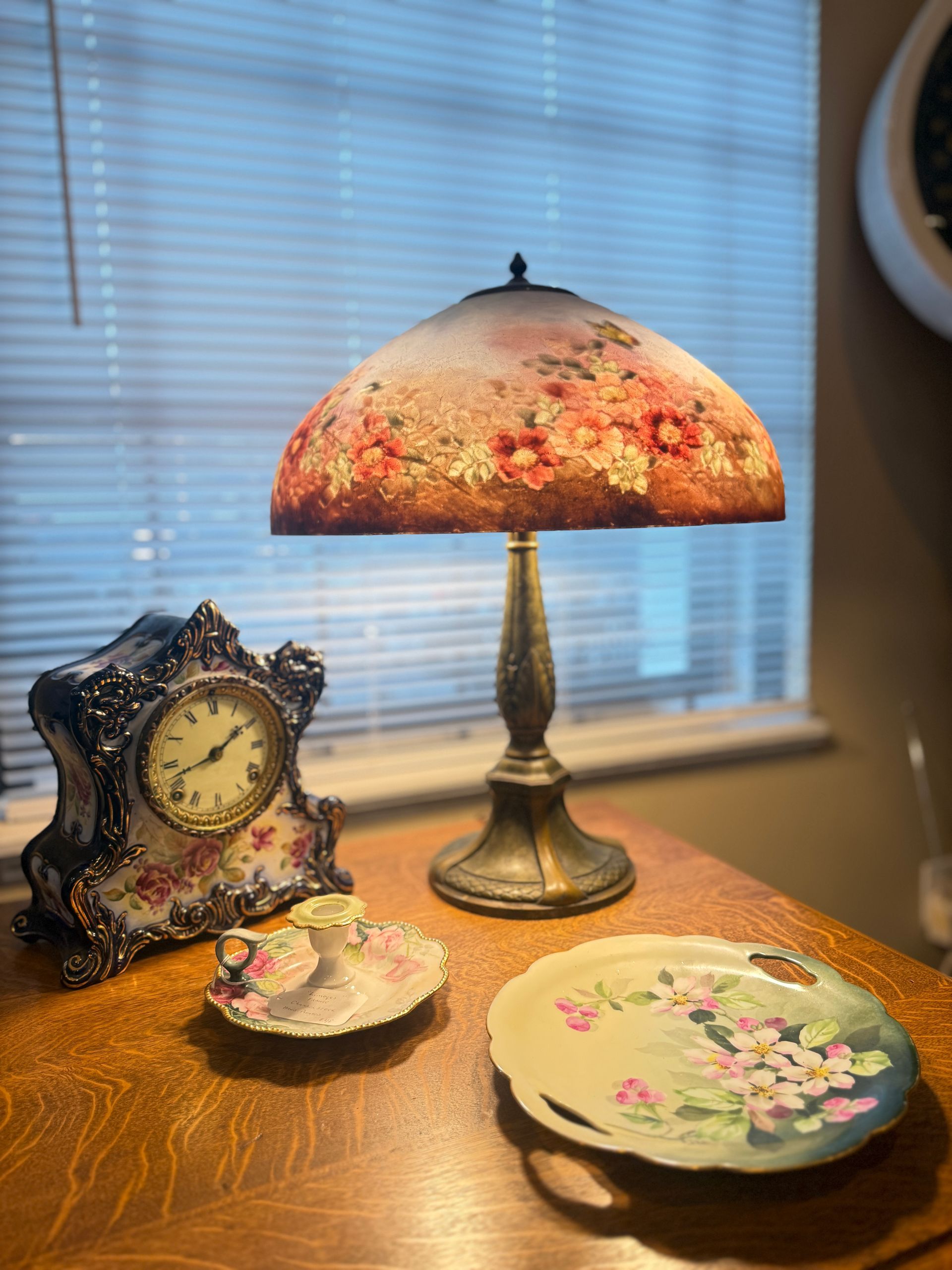 Table with floral lamp, clock, and plates in front of a window with blinds.