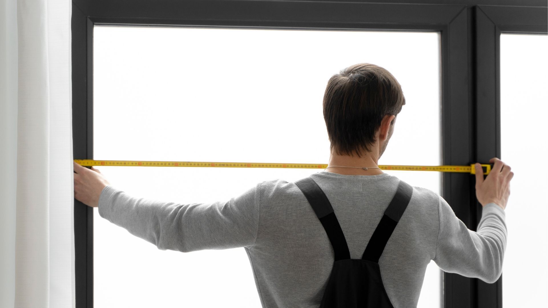 Person measuring a window frame with a tape measure, standing with arms outstretched.