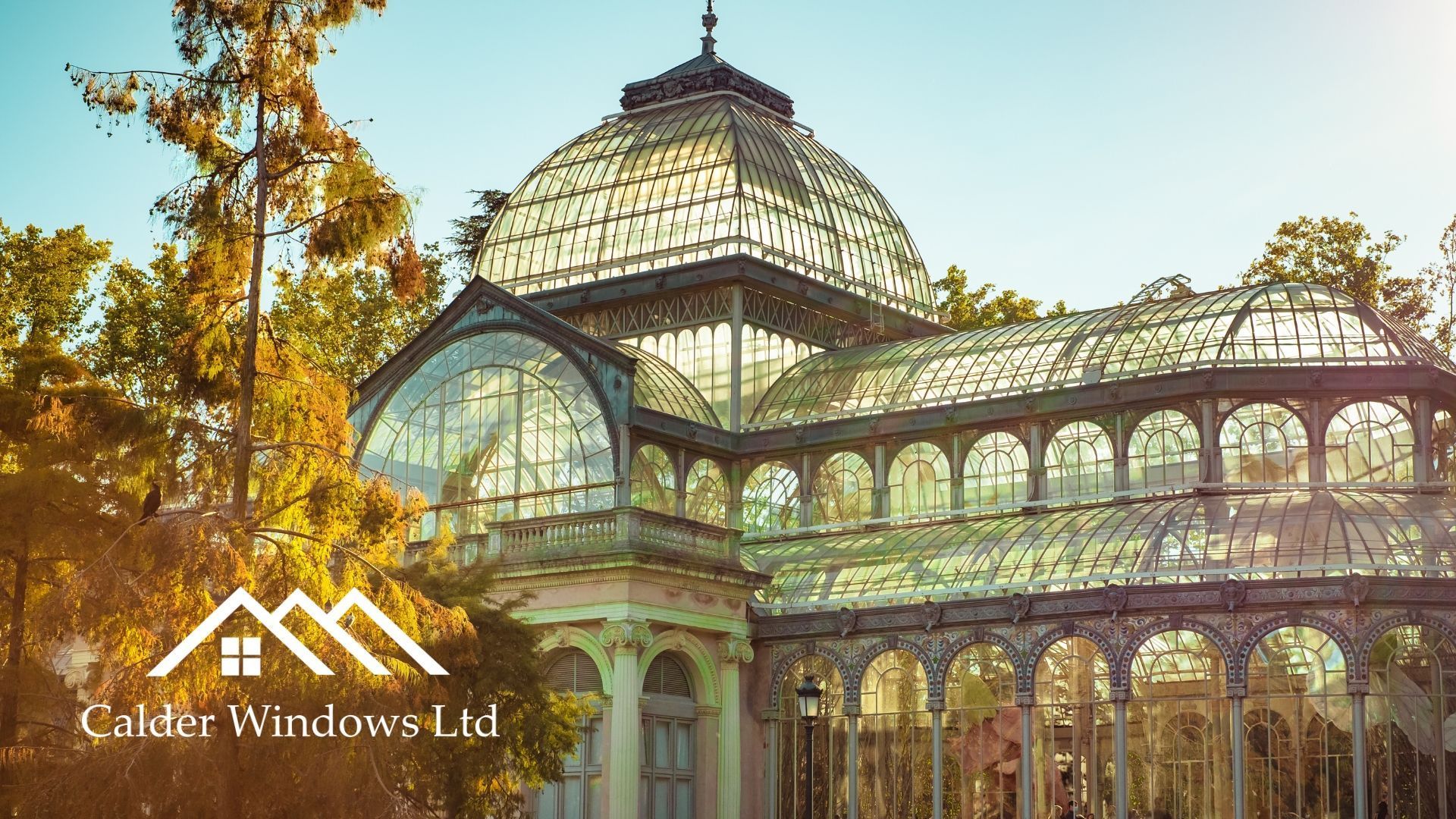 Ornate glass-and-iron conservatory with a domed roof in a sunlit autumn garden, with El Escorial logo watermark