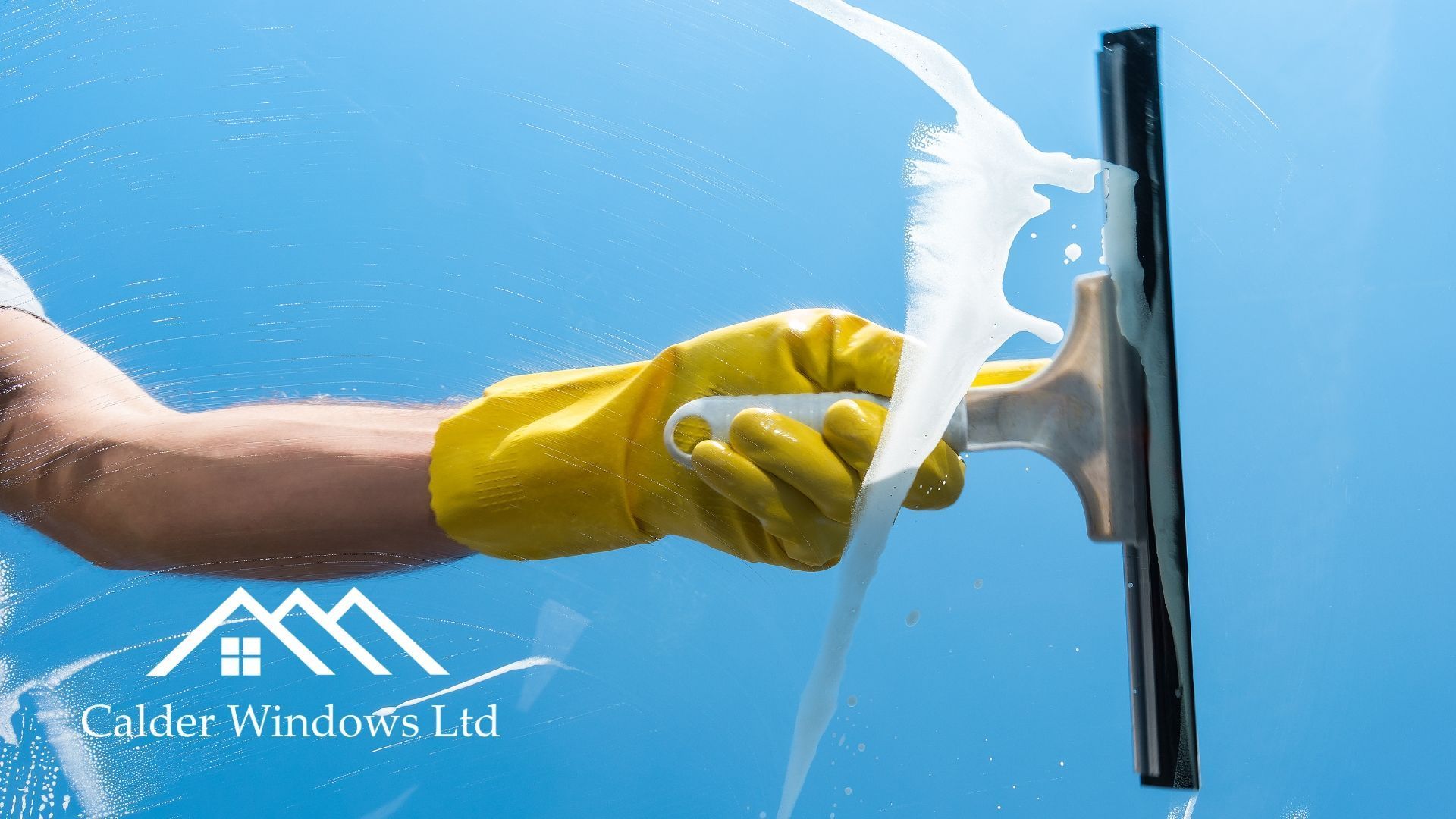 Gloved hand using a squeegee to wipe soap from a glass window against a blue sky.