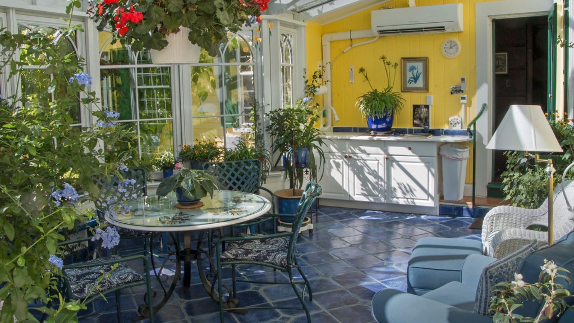 Sunny courtyard patio with round table, metal chairs, potted plants, and a yellow house facade with a serving counter