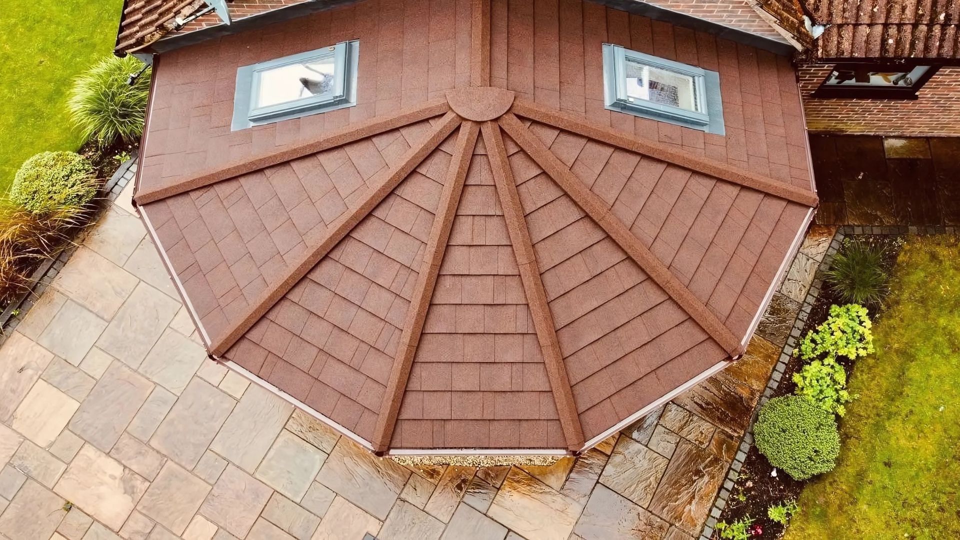 Aerial view of a brown shingled roof with two skylights and a stone patio below.
