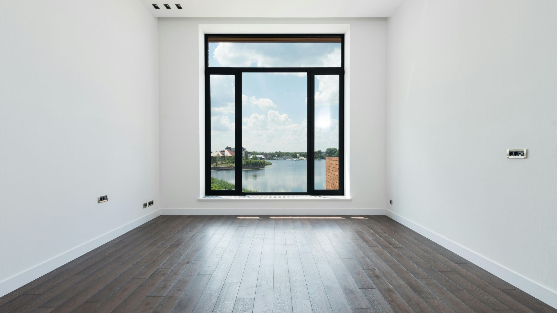 Empty modern room with dark wood floor and large window overlooking water and sky