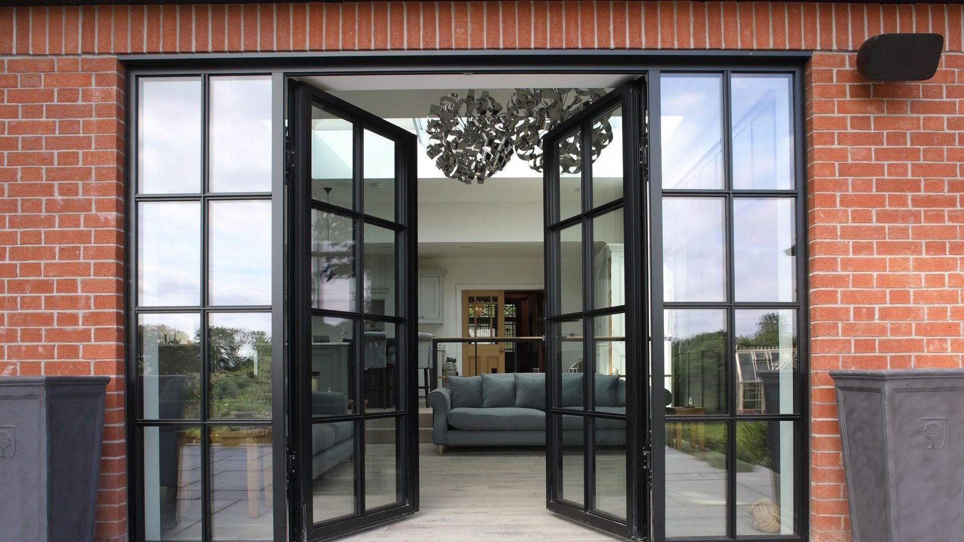 Open black French doors in a brick wall reveal a modern living room with a gray sofa.