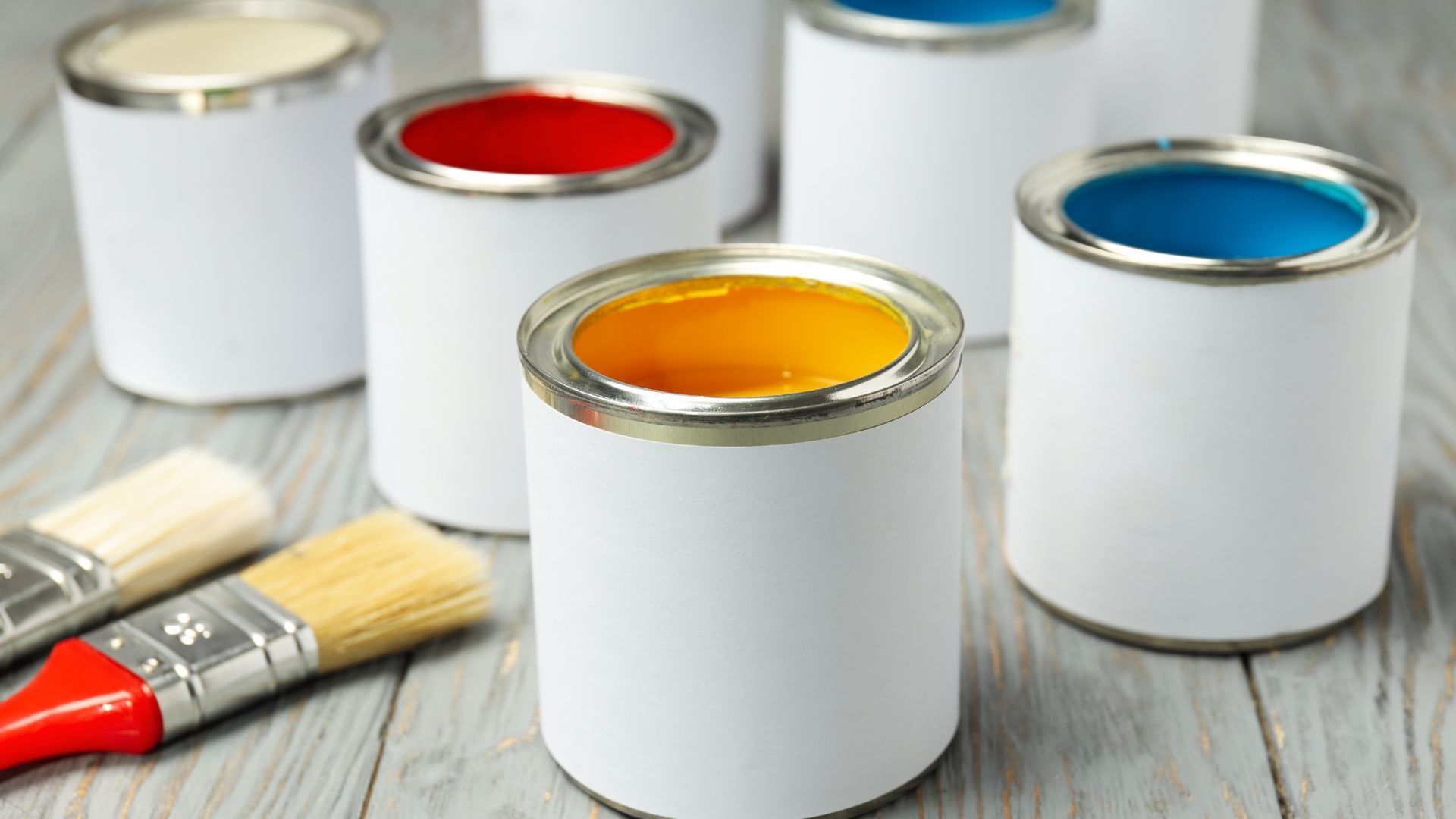 Paint cans with red, yellow, and blue paint beside brushes on a wooden surface