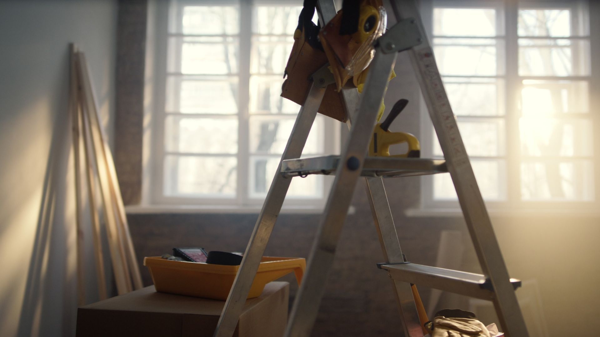 Sunlit room with a ladder beside a window and scattered tools, suggesting ongoing home repairs or painting