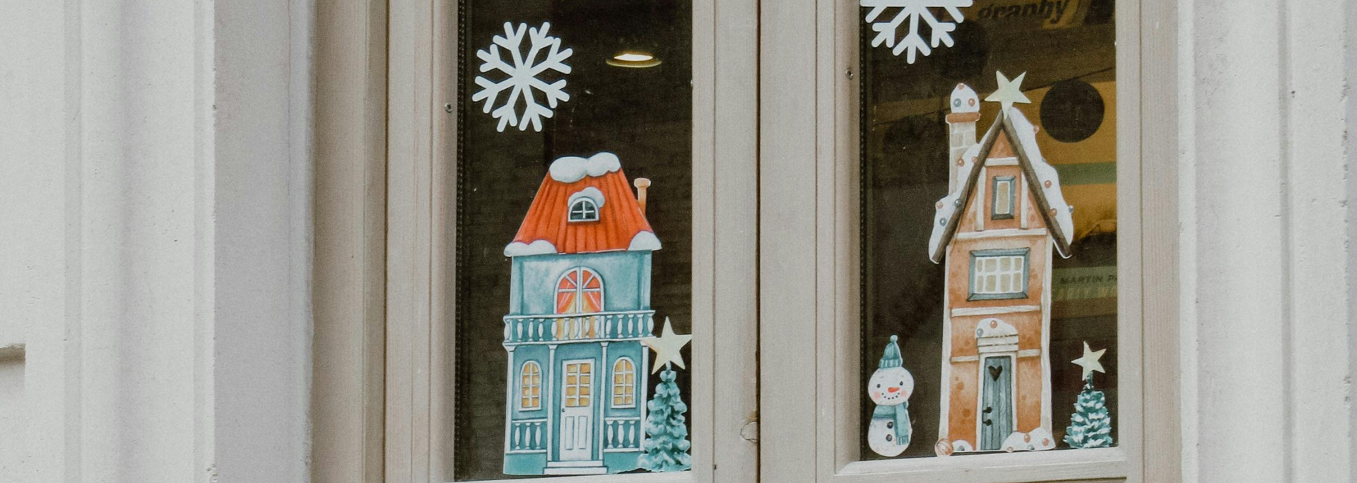 Two colorful house decorations displayed in window panes with white snowflake cutouts