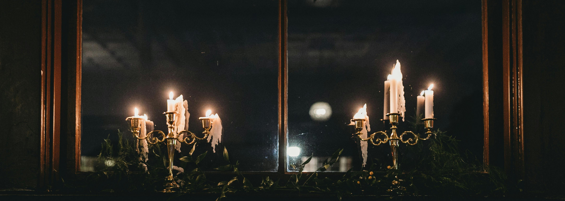 Candles glow on a dark table before a window at night, with reflections and a soft orange light.