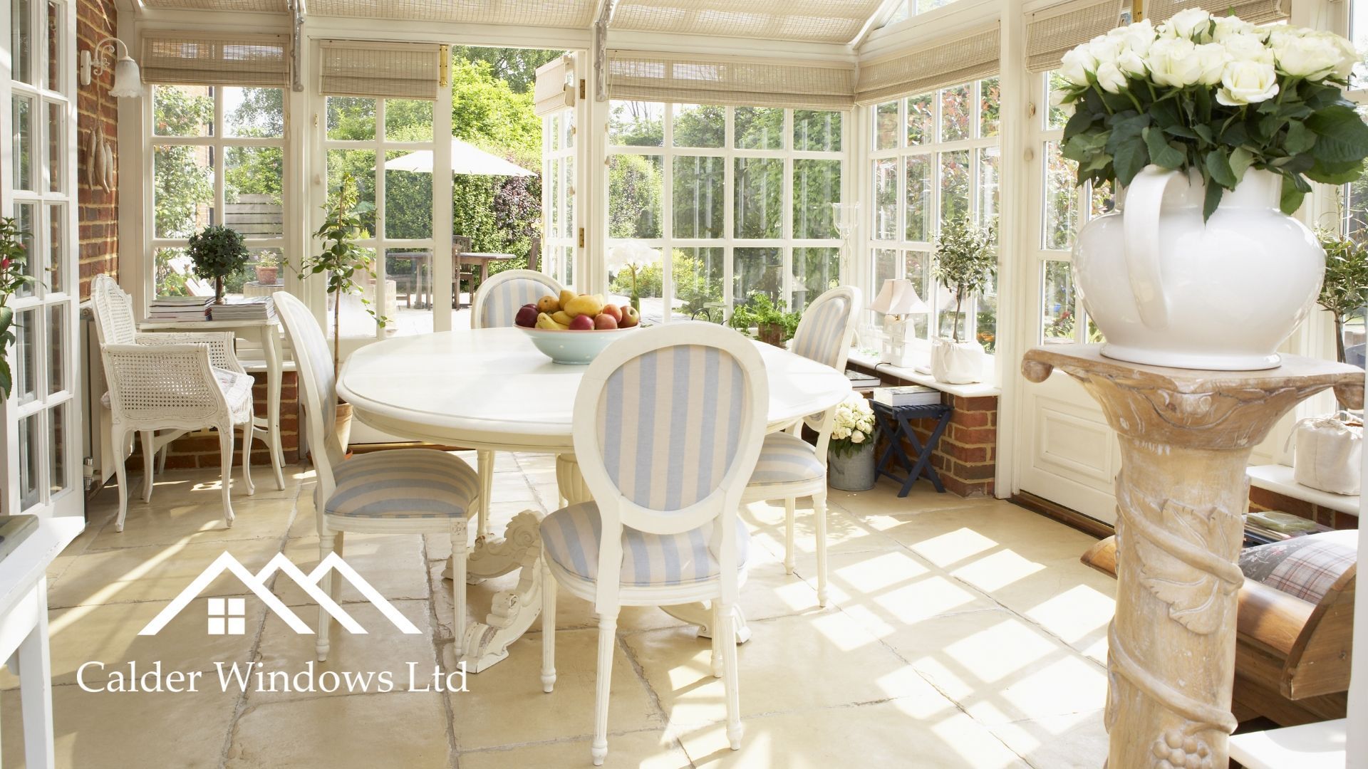 Bright sunroom dining area with white table, chairs, potted flowers, and large windows