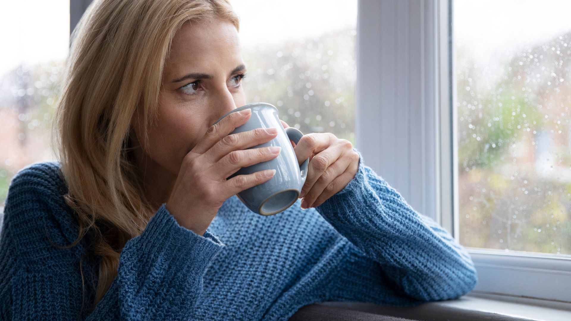 Woman in a blue sweater holding a mug by a window, looking outside.