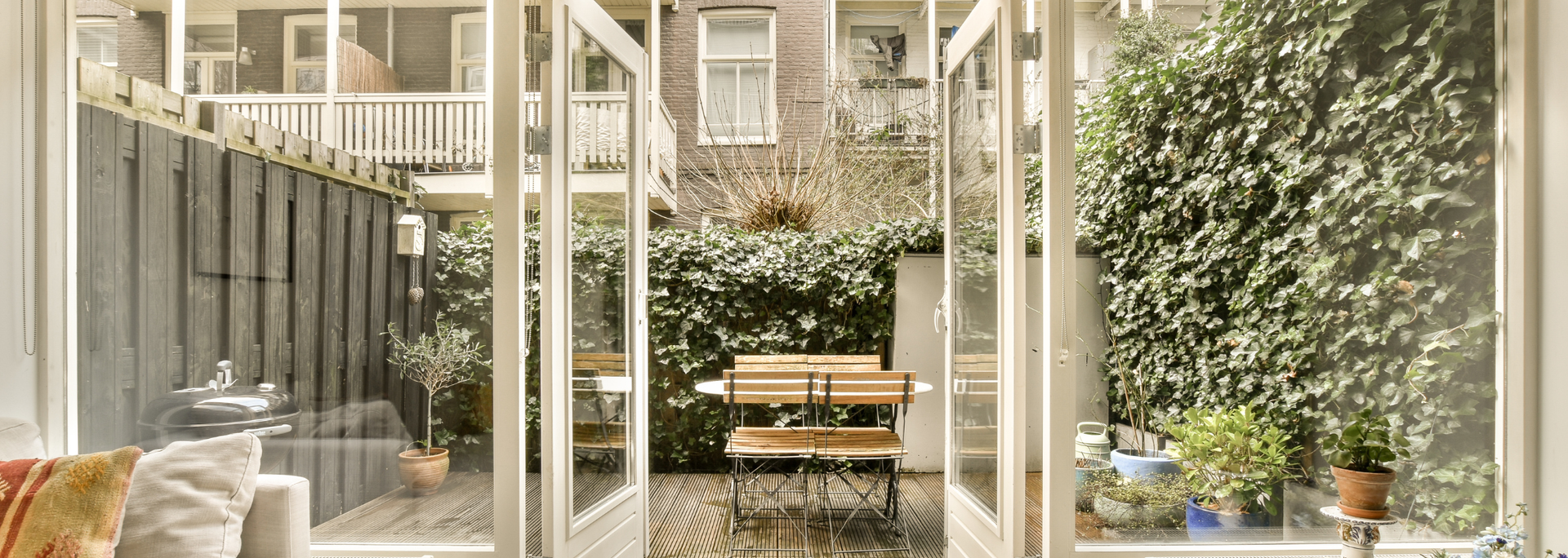 Small patio with wooden table and chairs, potted plants, and open glass doors in a sunlit courtyard.