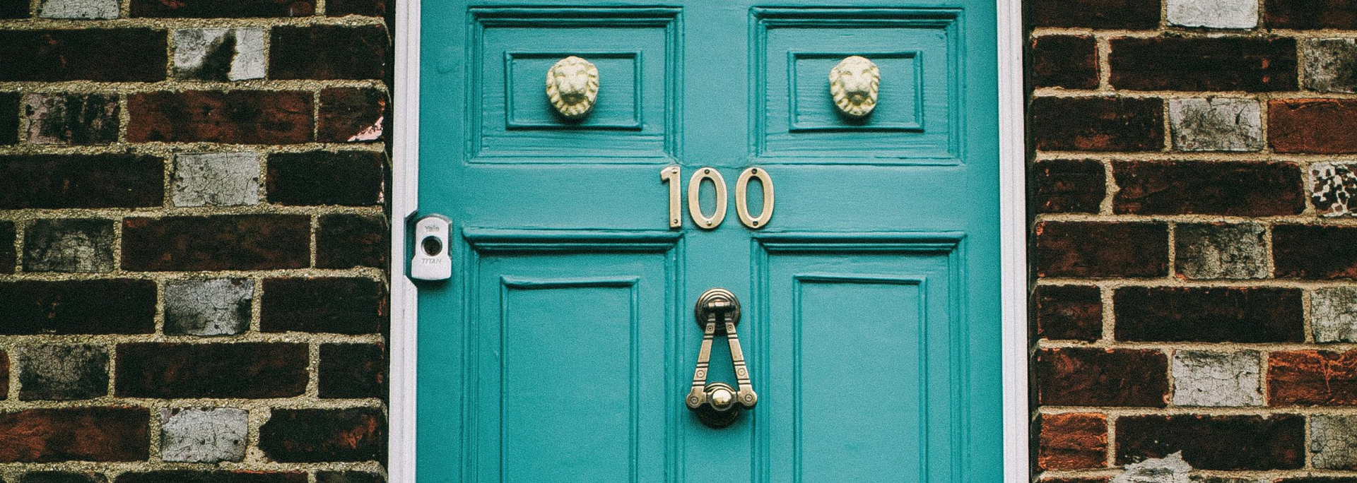 Teal front door with the number 100, brass knocker, and brick wall surround