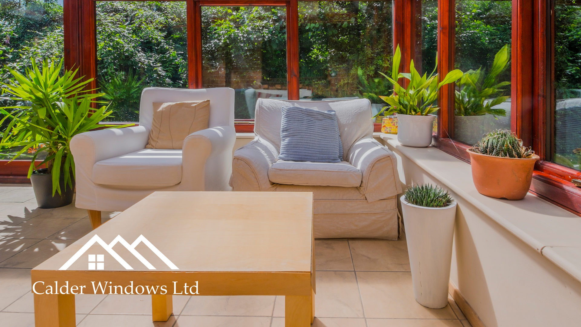 Bright sunroom with cream sofas, wooden table, and potted plants beside large windows