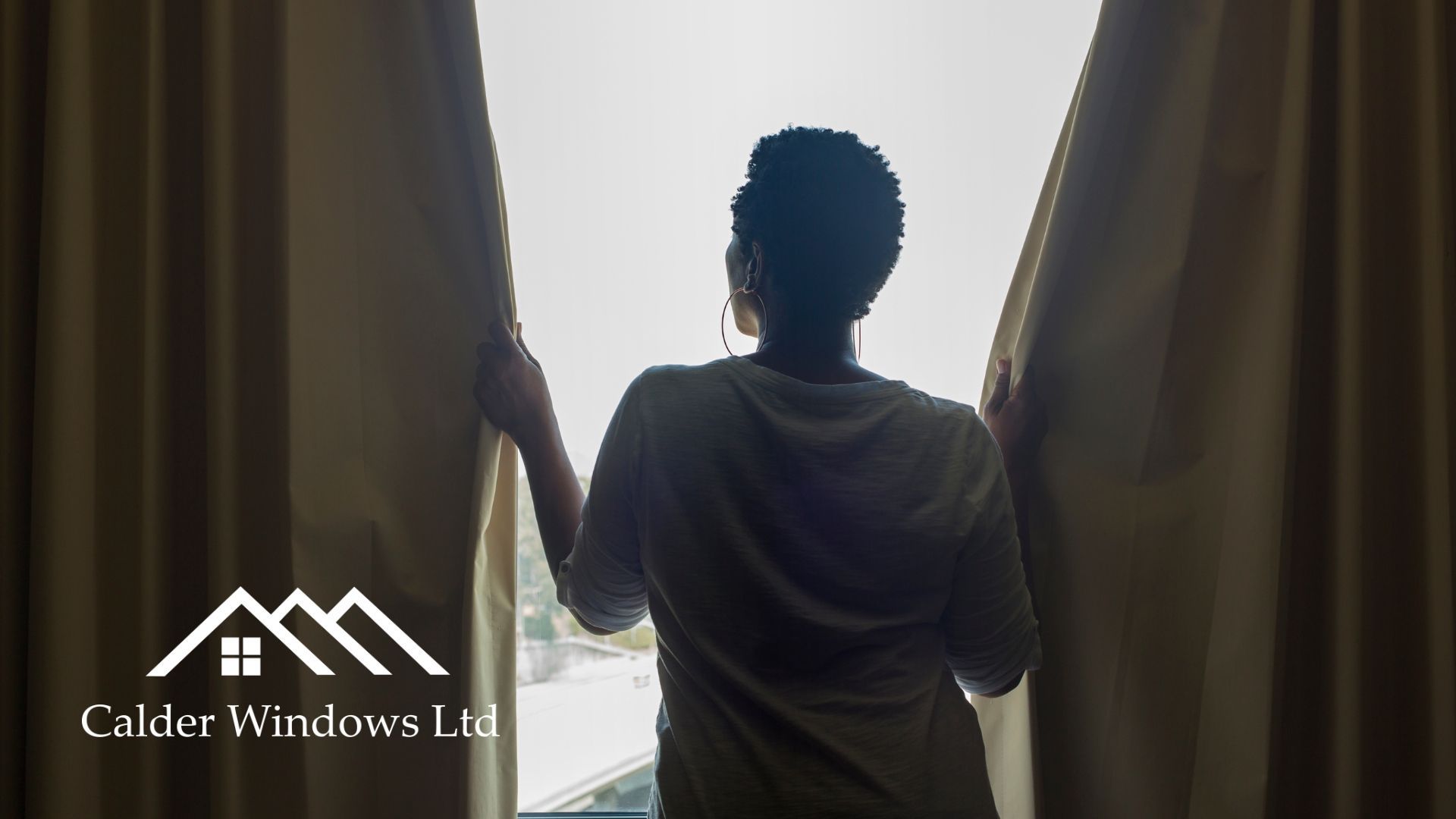 Silhouette of a person standing between open curtains, looking out a bright window.