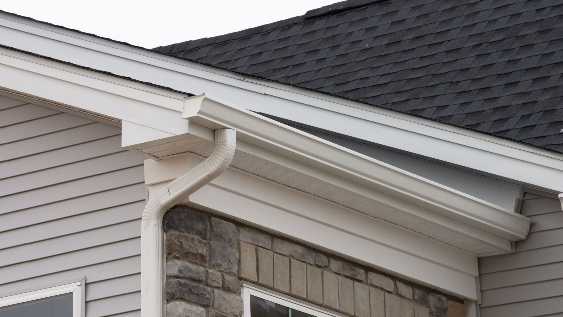 Close-up of a house roof edge with white gutter and downspout beside gray siding and stone facade