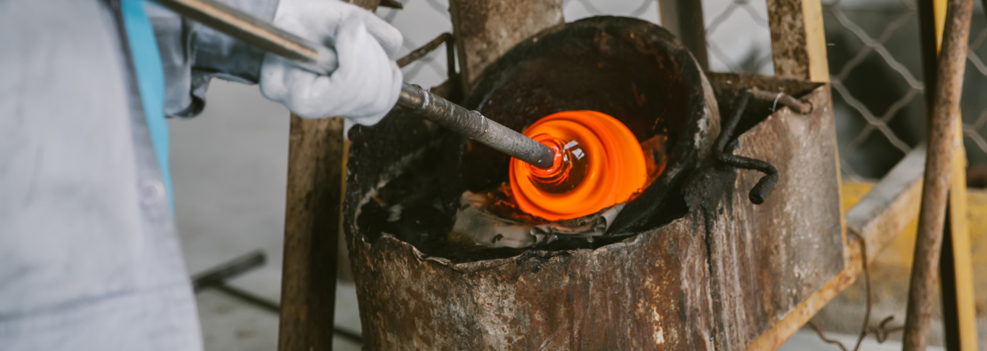 Gloved worker shaping glowing molten metal in a furnace with a long tool