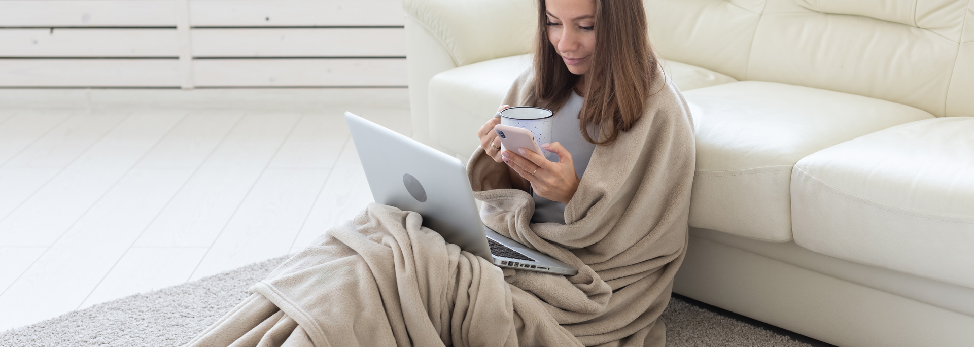 Woman using a laptop and smartphone on a white couch, wrapped in a beige blanket