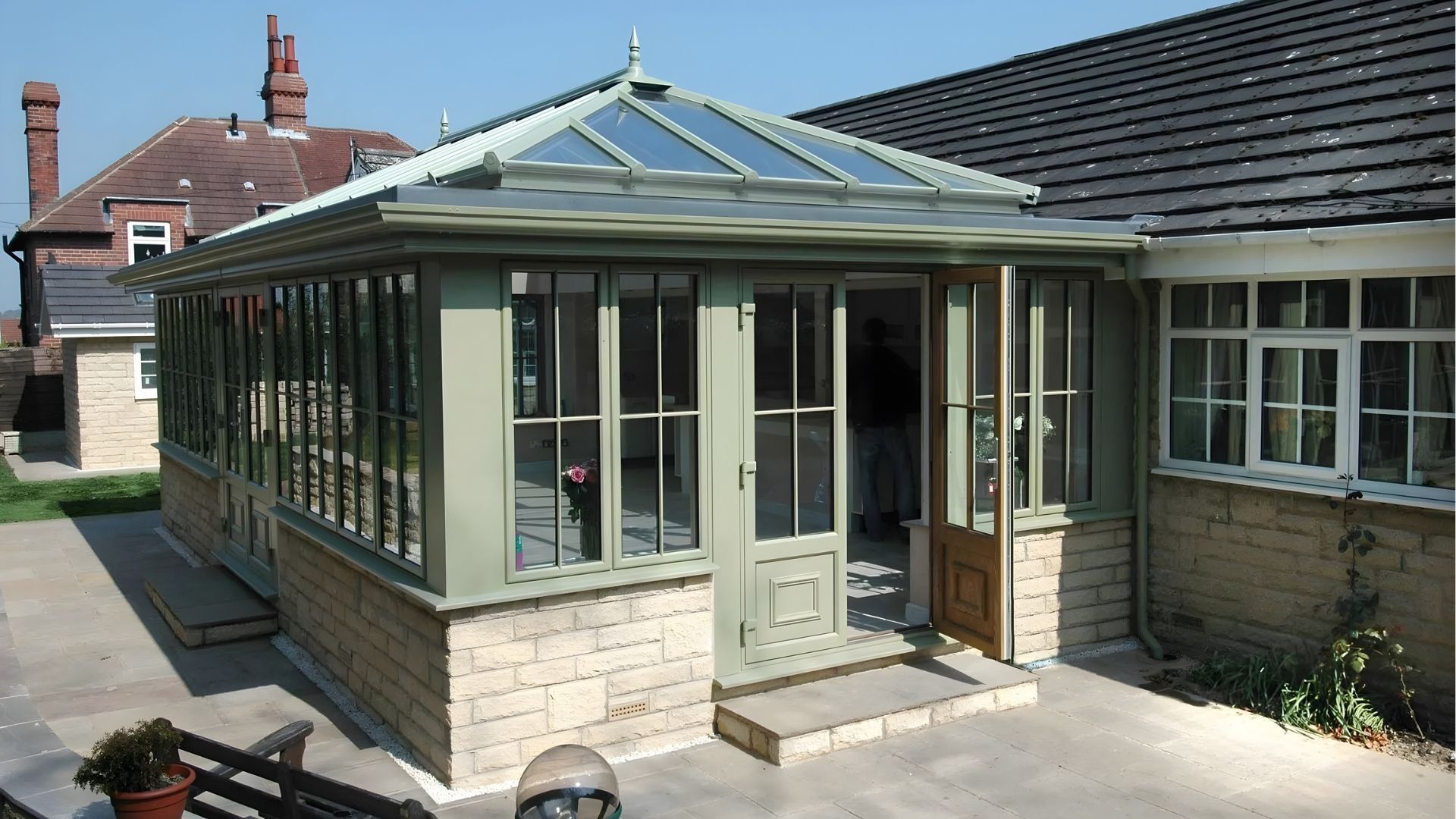 Glass sunroom with green trim attached to a stone house, viewed from the patio.