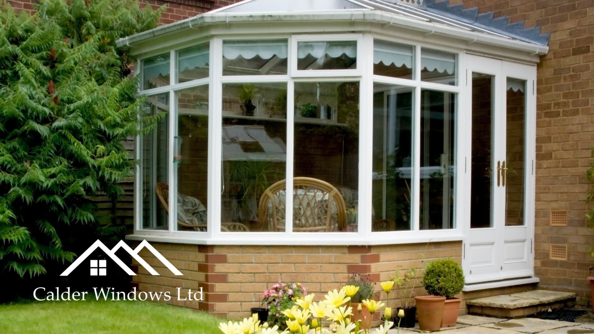 White conservatory with large windows attached to a brick house, surrounded by garden plants.