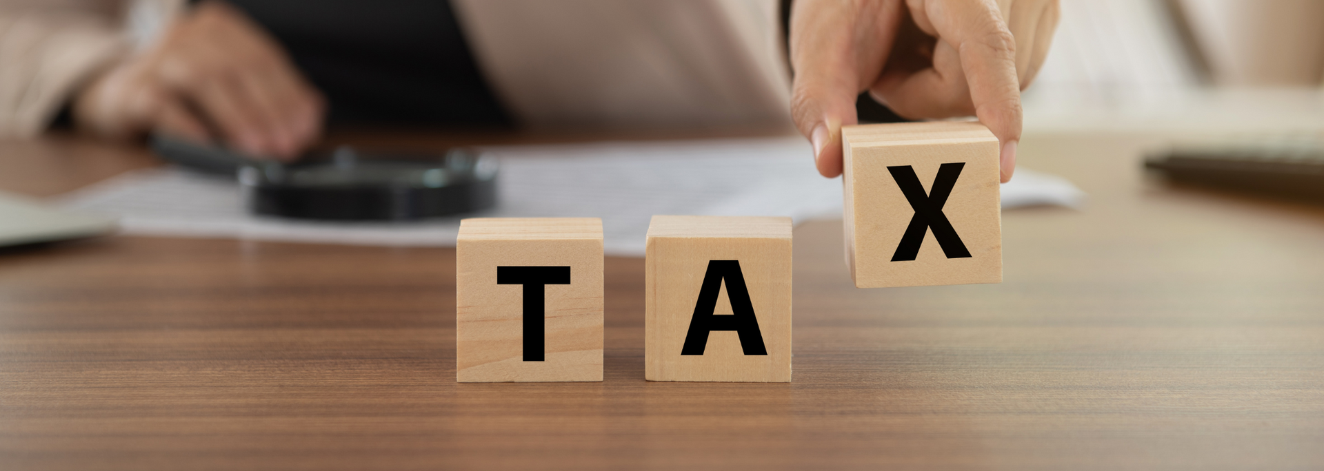 Wooden blocks spelling “TAX” on a desk, with a hand arranging the X block.