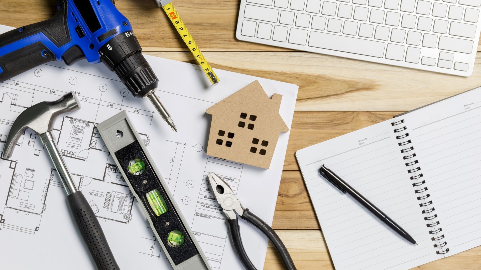 Workshop tools, pencil, notebook, and wooden house model on a desk with graph paper and keyboard.