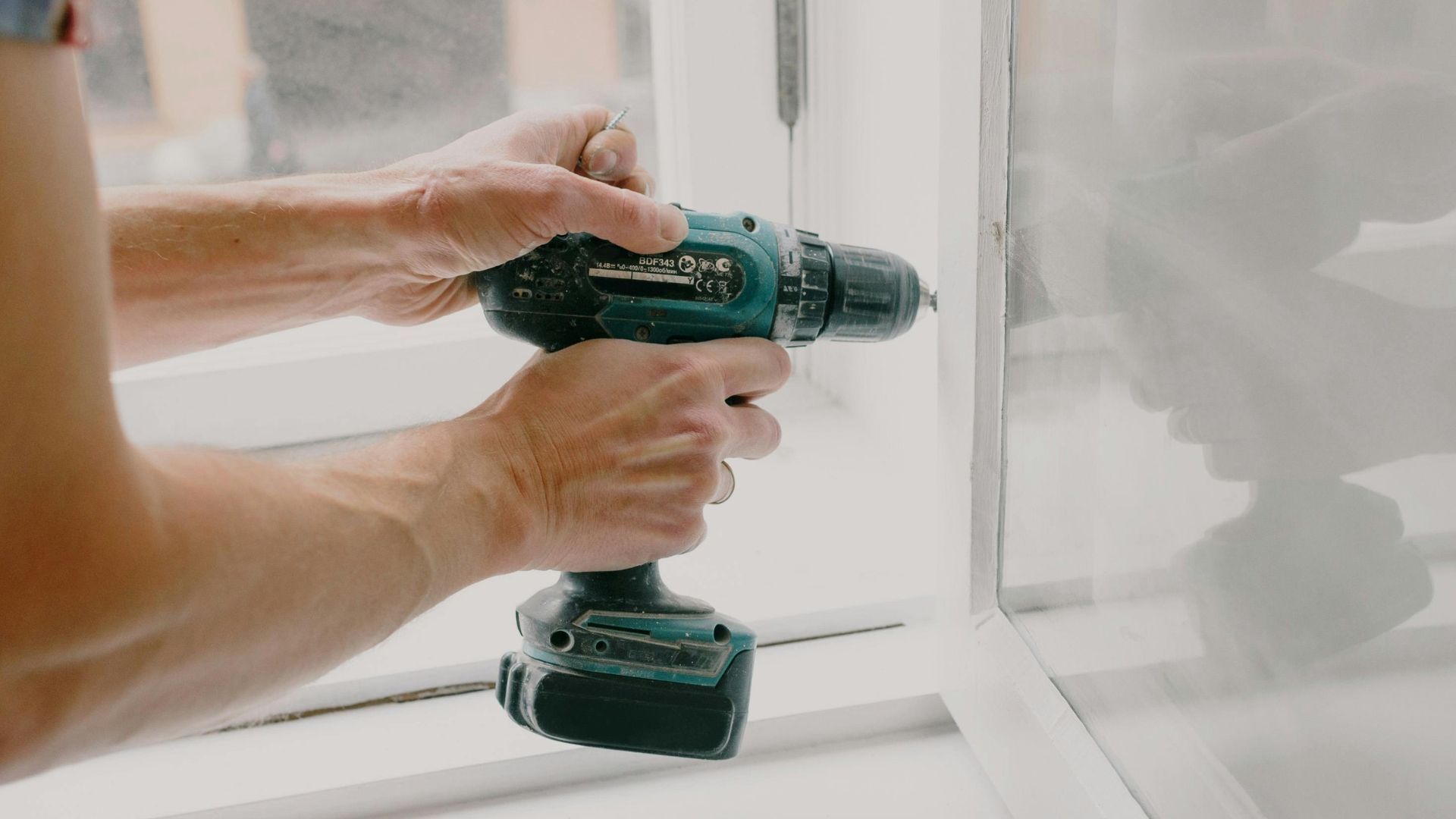 Hands using a green cordless drill on a white window frame during home repair