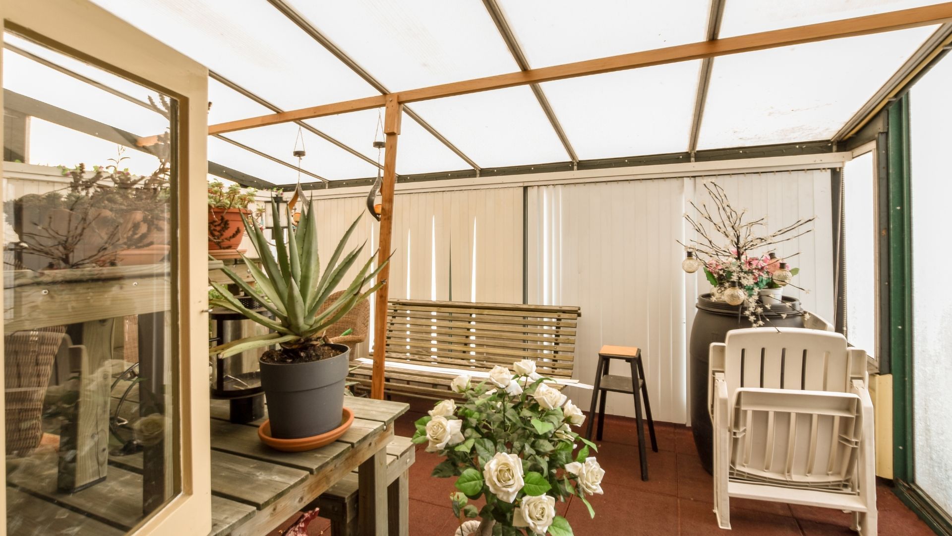 Bright sunroom with potted flowers, a wooden bench, white chair, and glass shelves on a tiled floor