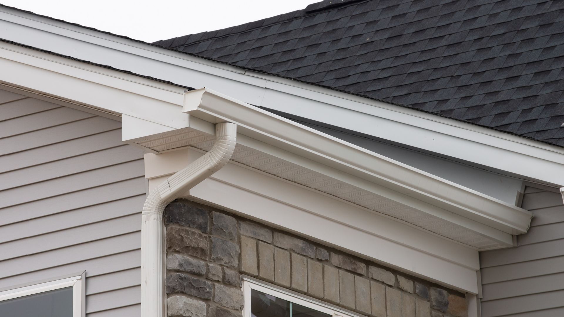 Close-up of a house roof eave with white gutters and beige siding under dark shingles