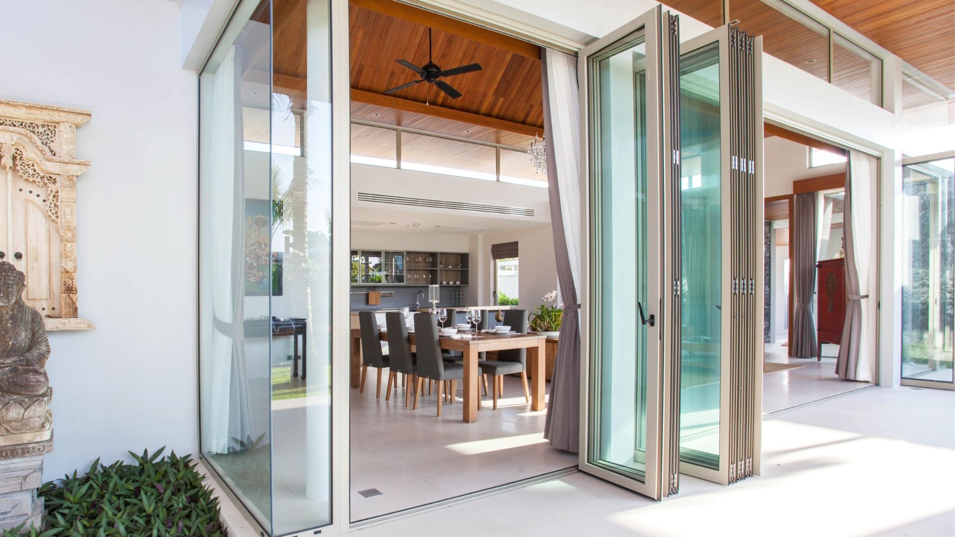 Open folding glass doors to a bright dining room with a wooden table and chairs, opening to a patio.