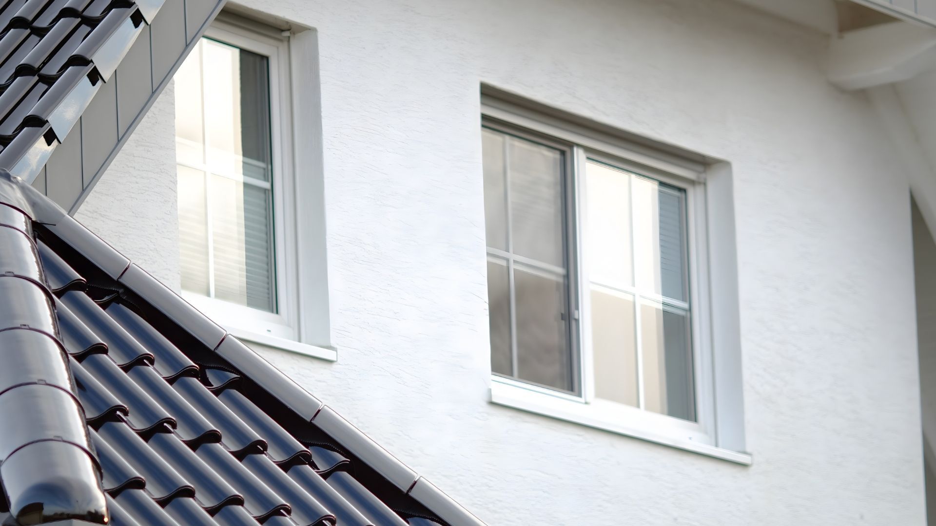 White house wall with two windows beside a dark tiled roof and a staircase railing