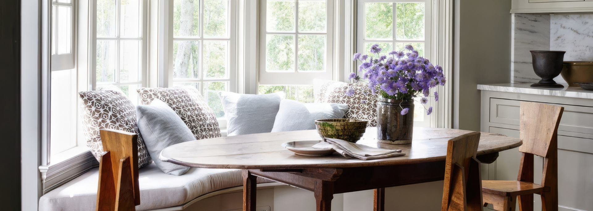 Bright living room with a wooden table, patterned chairs, flowers, and a desk by large windows.