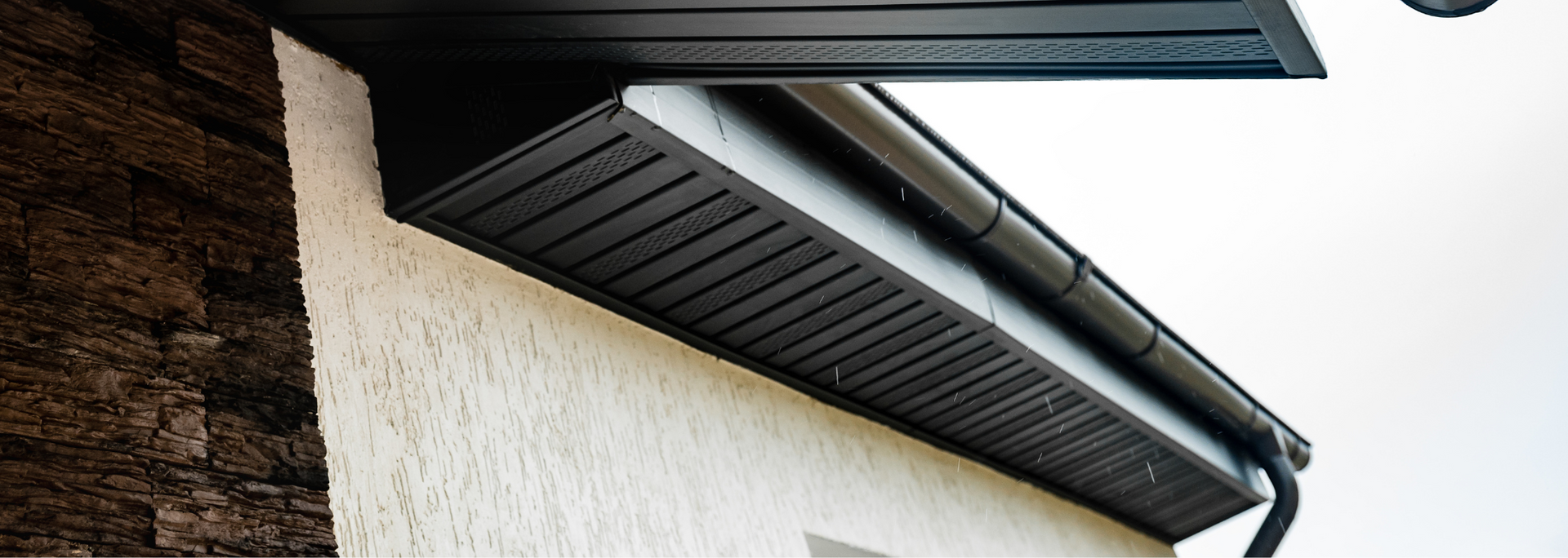 Close-up of a house roof eave with dark gutters and soffit against white siding and brick wall