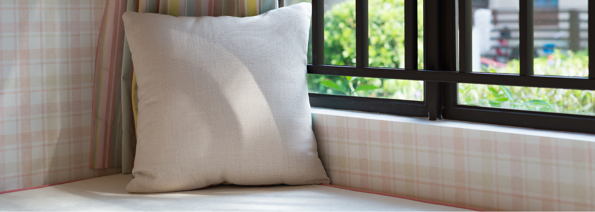 Beige cushion on a windowsill beside plaid curtains, with a window view of greenery.