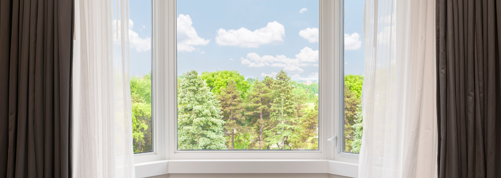 Window with white curtains overlooking green trees and a cloudy sky
