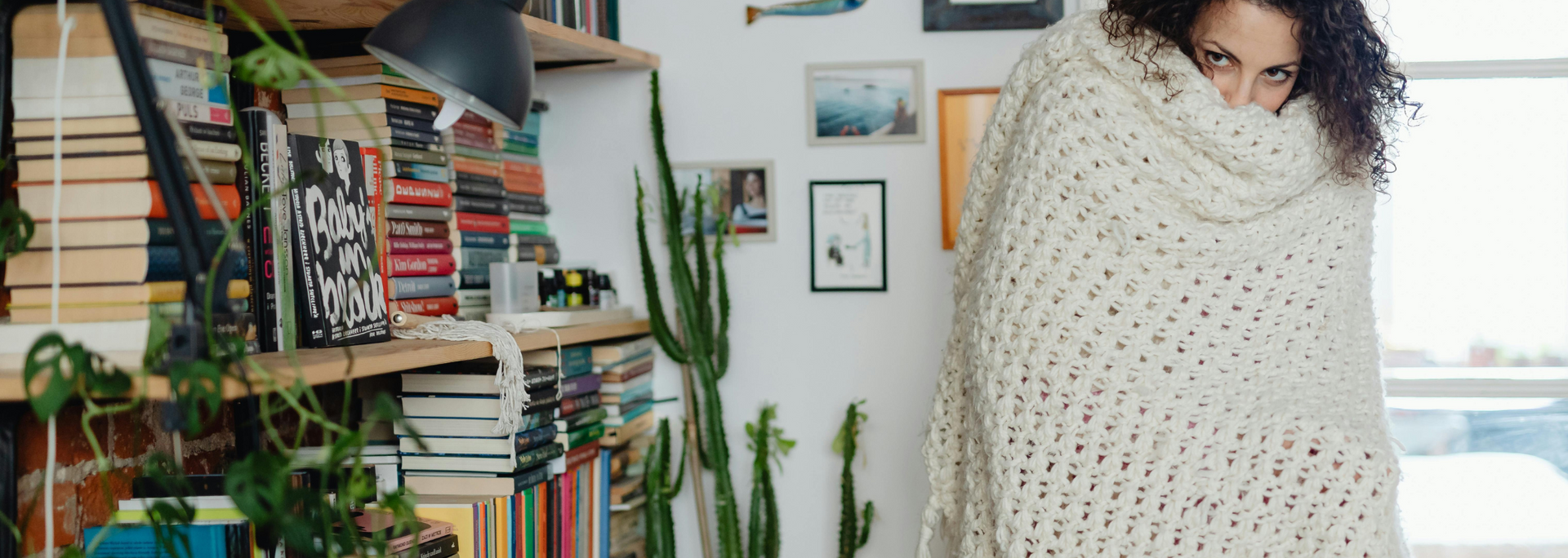 Person wrapped in a cream knit blanket beside a bookshelf and potted plants, in a cozy room