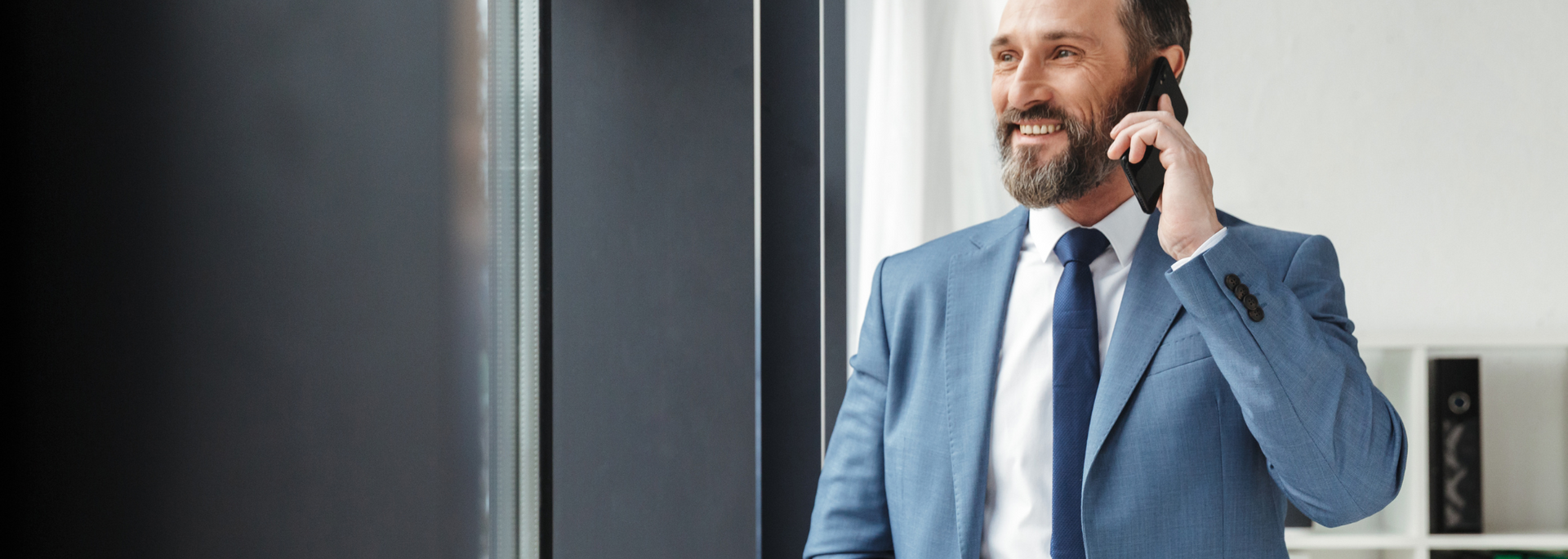 Man in a blue suit talking on a phone near a glass wall in an office hallway