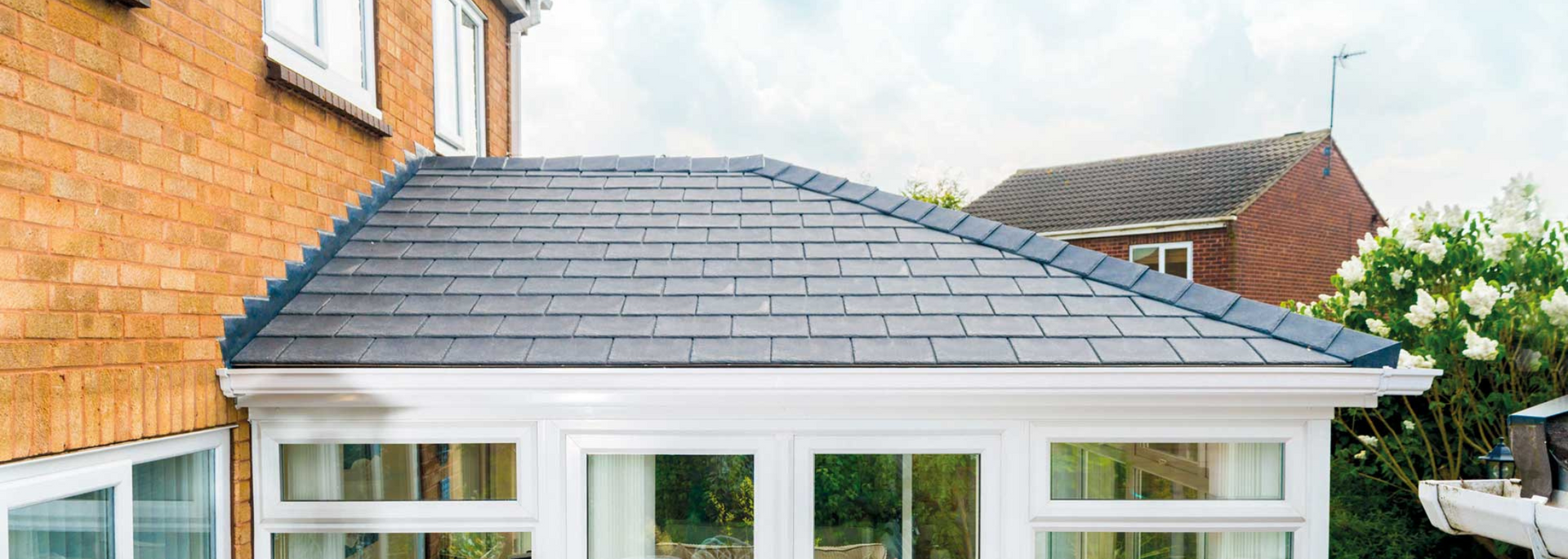 Backyard conservatory with gray tiled roof and white-framed windows beside an orange brick house