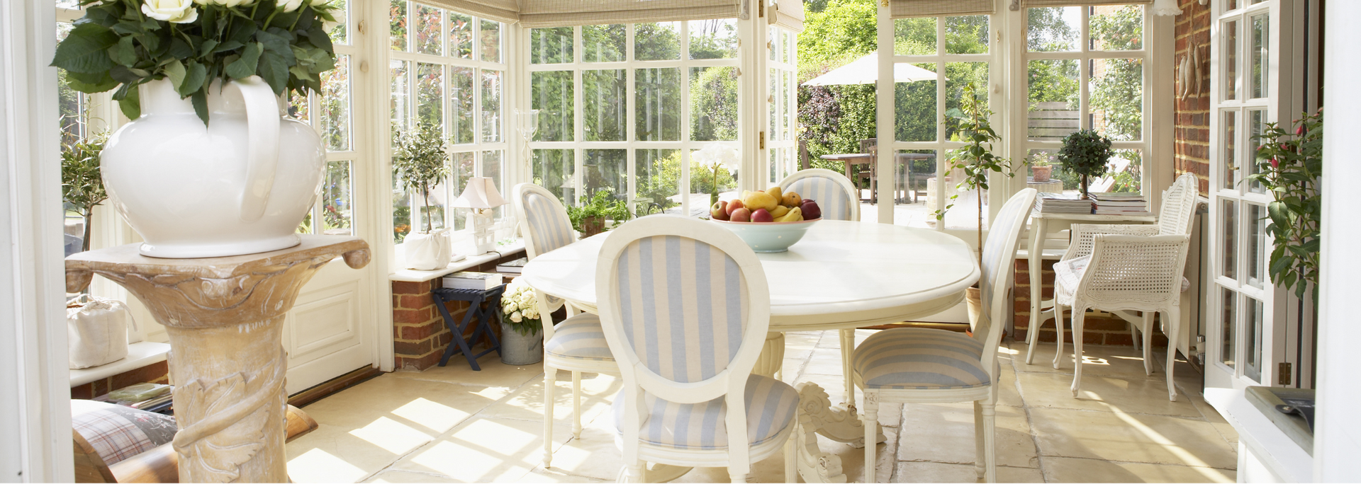 Bright sunroom with white wicker chairs, round table, and large windows overlooking a lush garden.