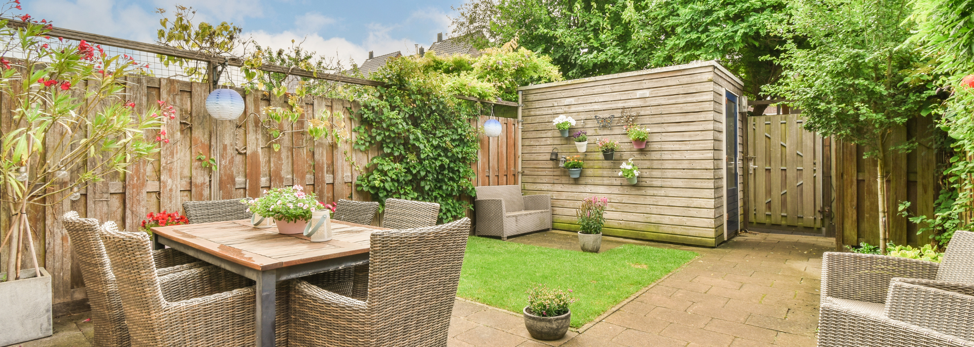 Backyard patio with wicker furniture, potted plants, and a wooden shed under a sunny sky