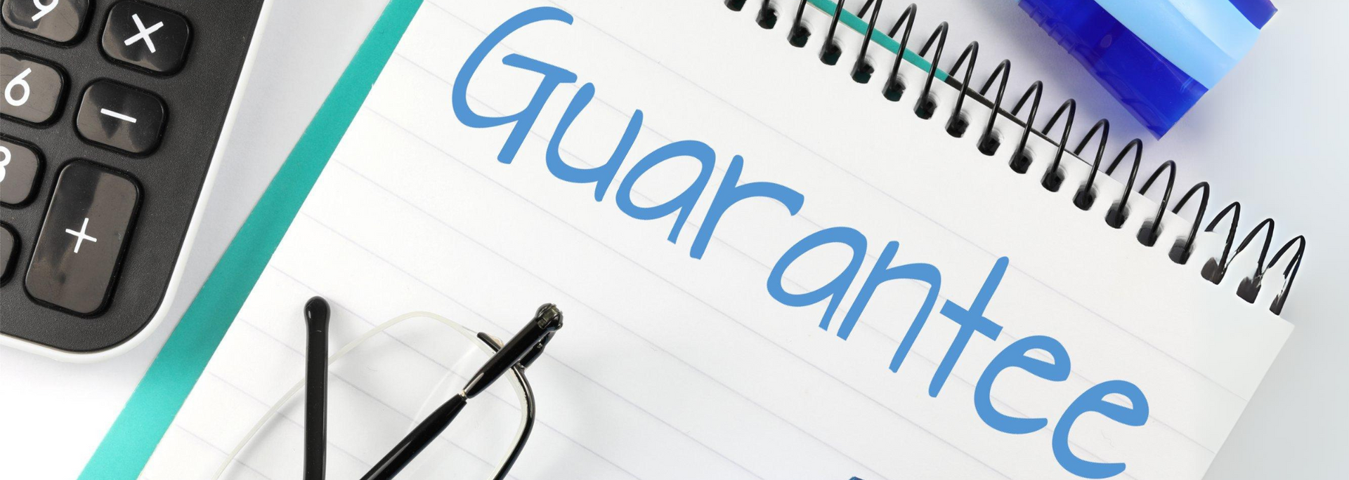 Notebook page with the word “Guarantee” handwritten in blue, beside eyeglasses and a calculator on a desk