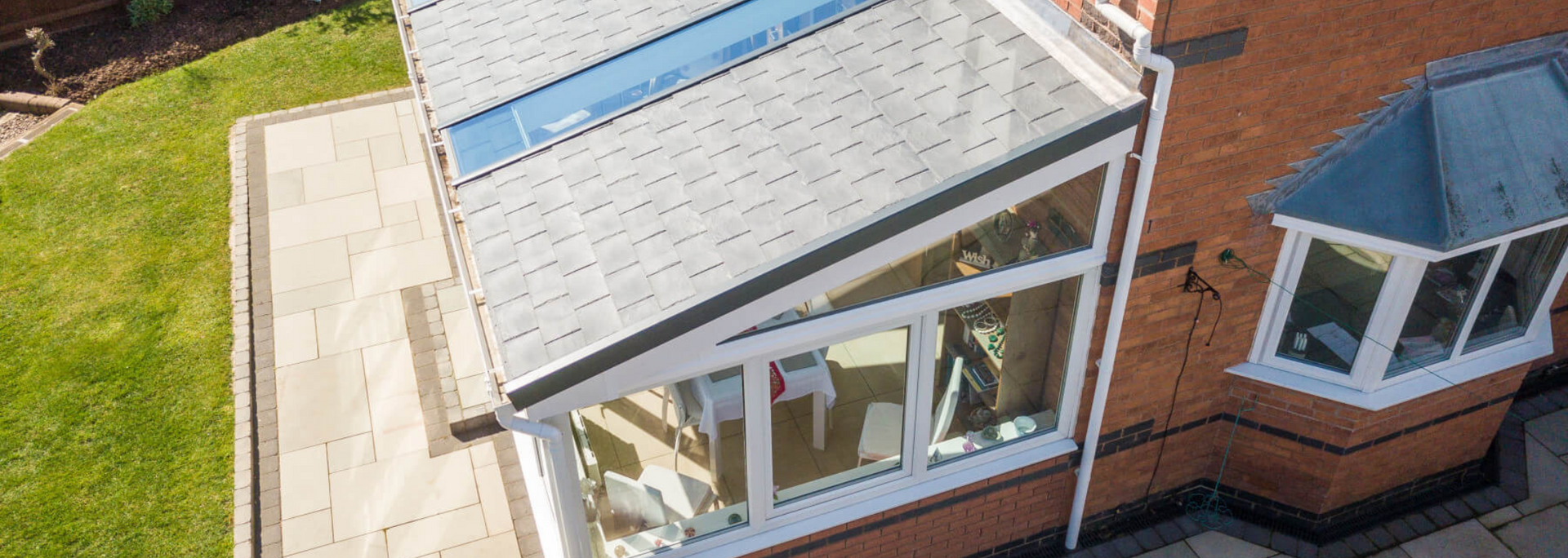 Aerial view of a house roof with a glass sunroom and patio beside a grassy yard.