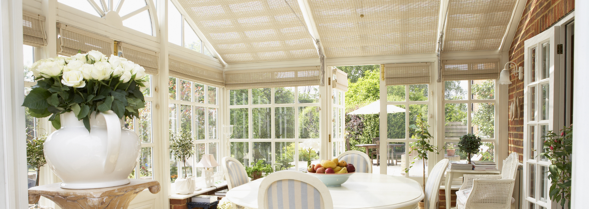 Bright sunroom with white furniture, glass walls, and a large floral arrangement on a table