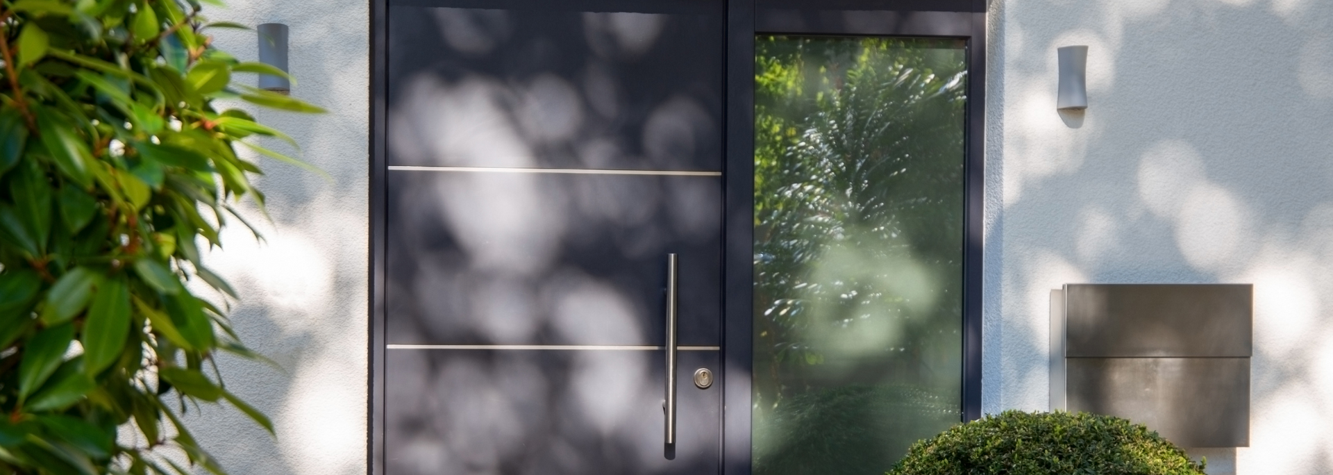 Modern house entrance with gray door, green glass door, and plants by a sunlit wall